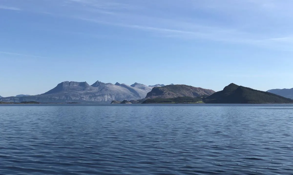 Når du kommer sydfra har du De Syv Søstre i bakgrunnen på vei inn mot Tro. Ankringsplassen ligger nedenfor de mørke og skogkledde toppene nærmest og til høyre i bildet